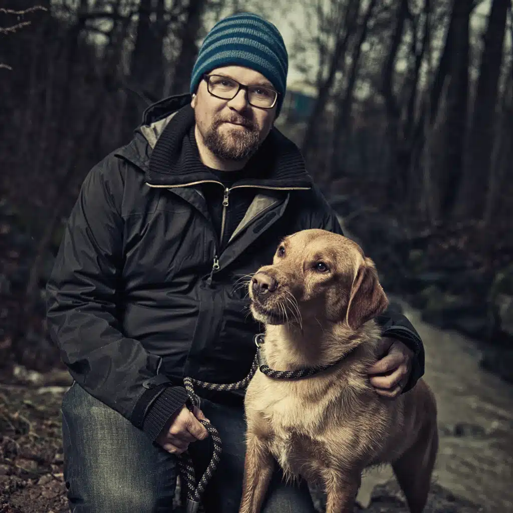 Mann mit Brille und Mütze sitzt im Wald neben seinem Hund während eines Spaziergangs